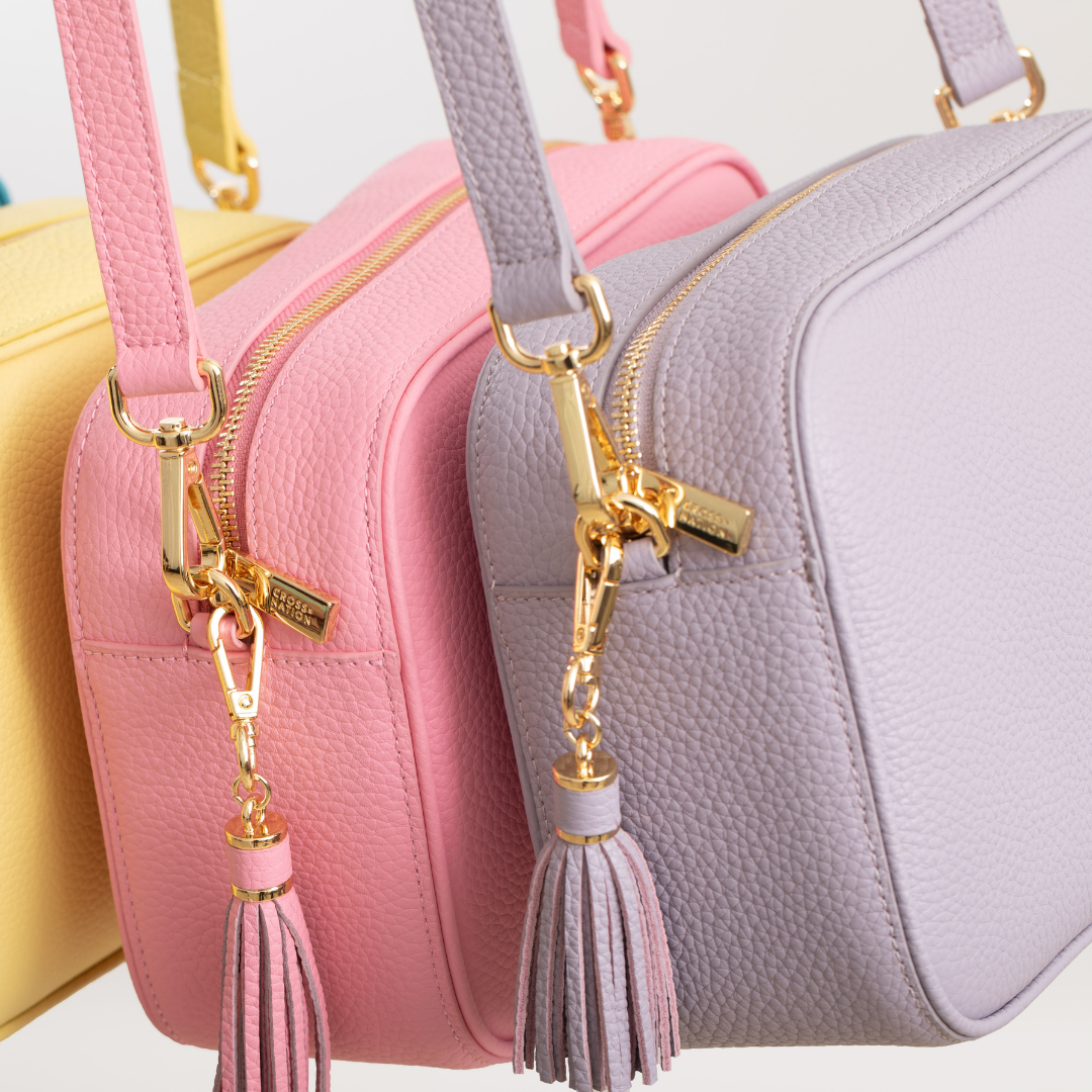 Three handbags in yellow, pink, and purple with matching tassel keychains on a light background.
