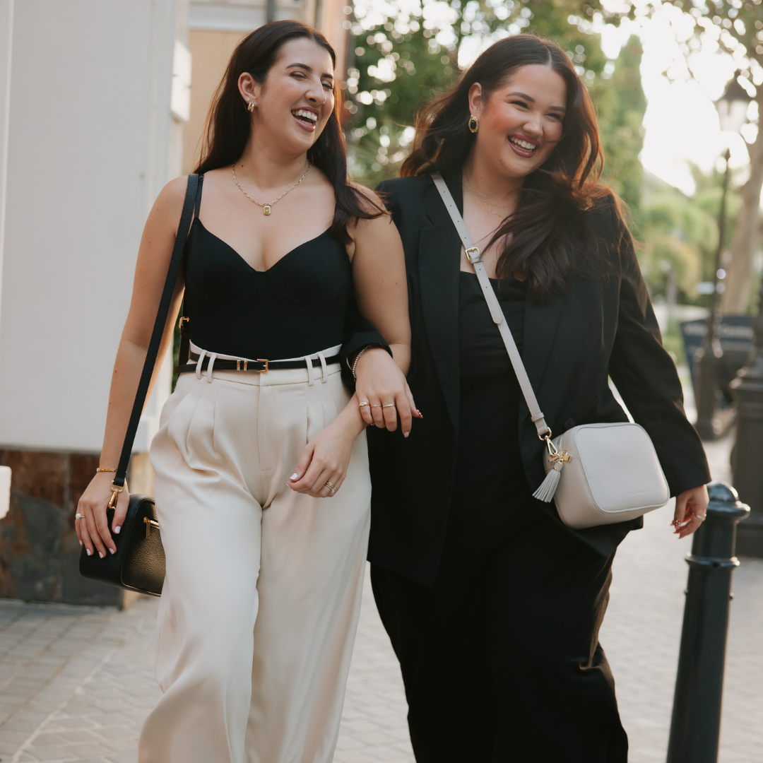 Two women walking together on a street, smiling wearing leather crossbody bags.