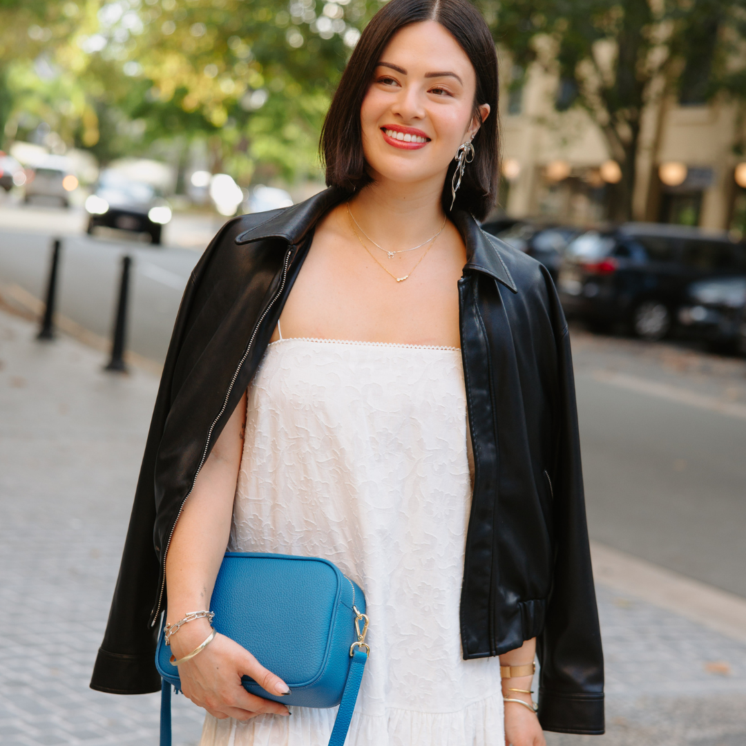 Woman in a white dress and black leather jacket holding a blue handbag on a city street.