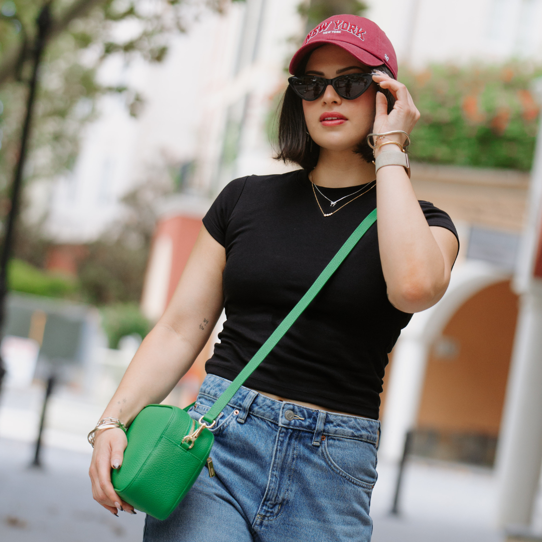 Woman wearing a black top, blue jeans, and a red cap, holding a green bag and a phone.