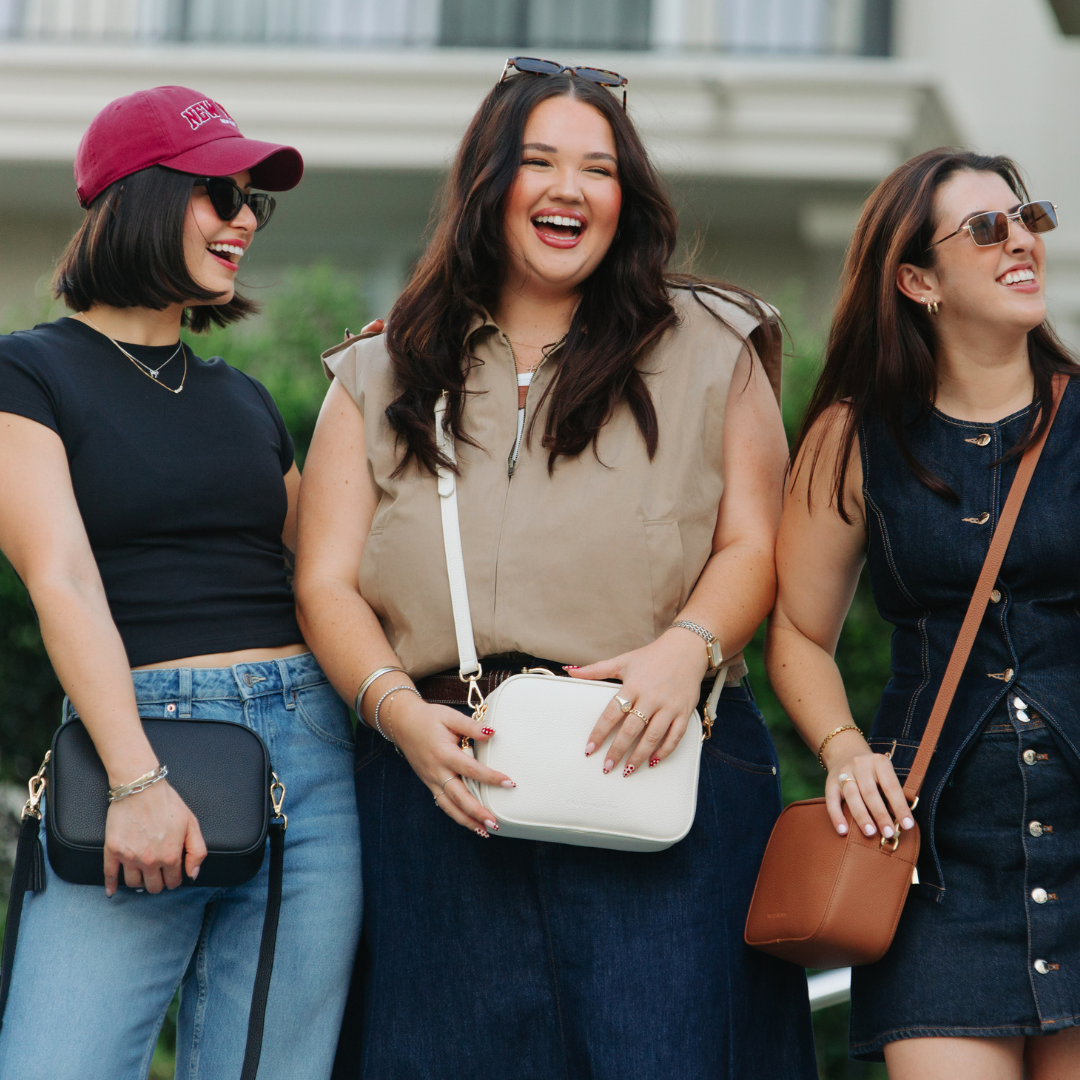 Three women standing together outdoors, smiling and holding handbags.