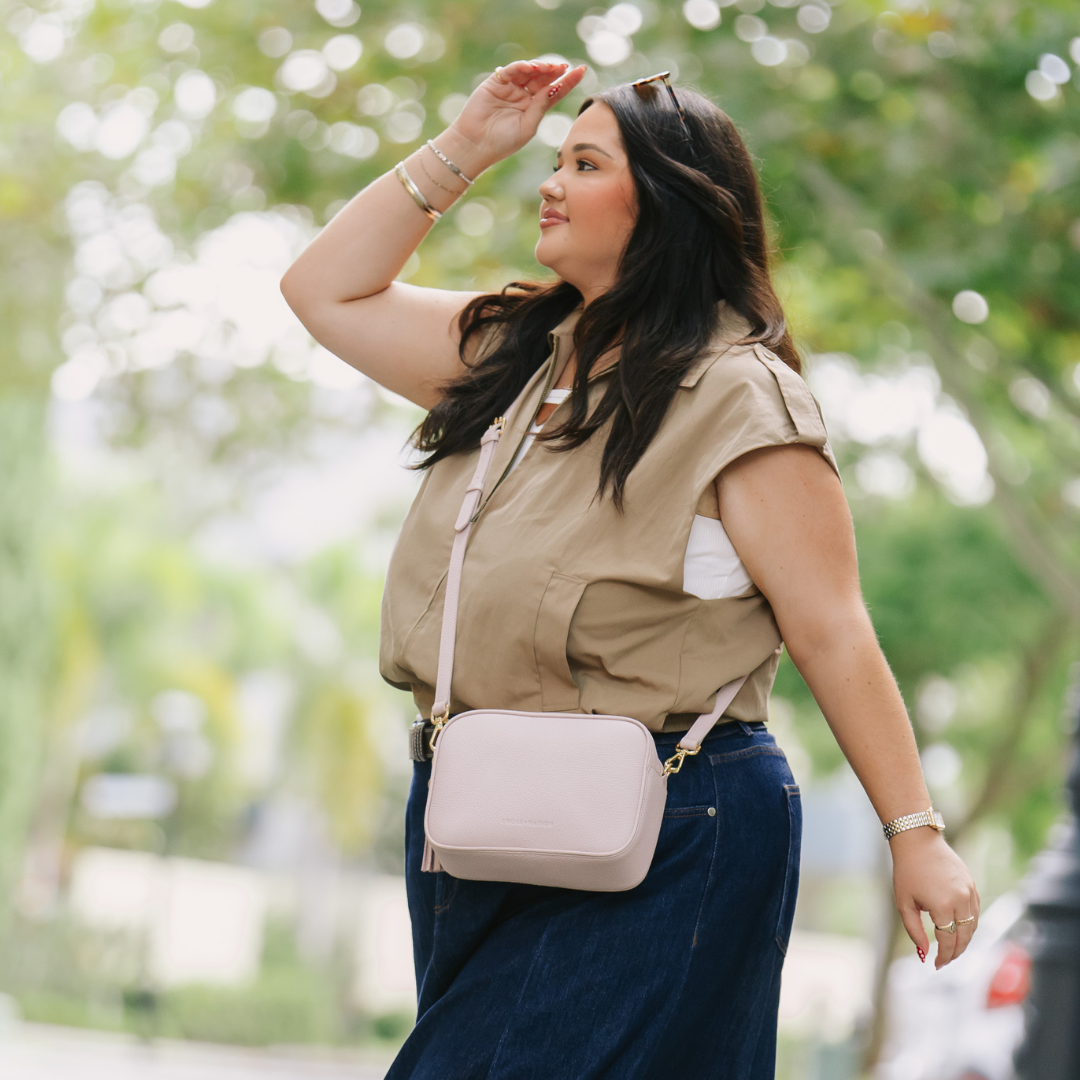 Woman walking outdoors with a pink crossbody bag, wearing a beige shirt and blue jeans.