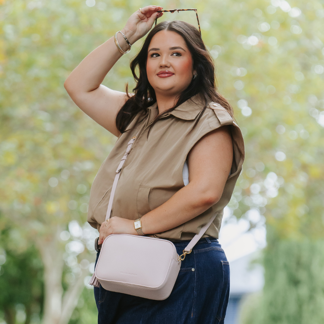 Woman holding a light pink crossbody bag outdoors with greenery in the background
