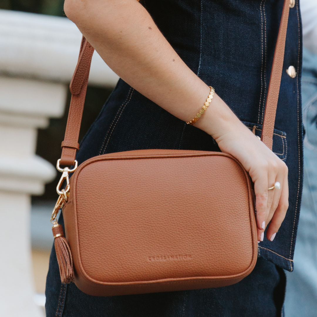 Brown leather crossbody bag held by a person wearing a denim jacket.