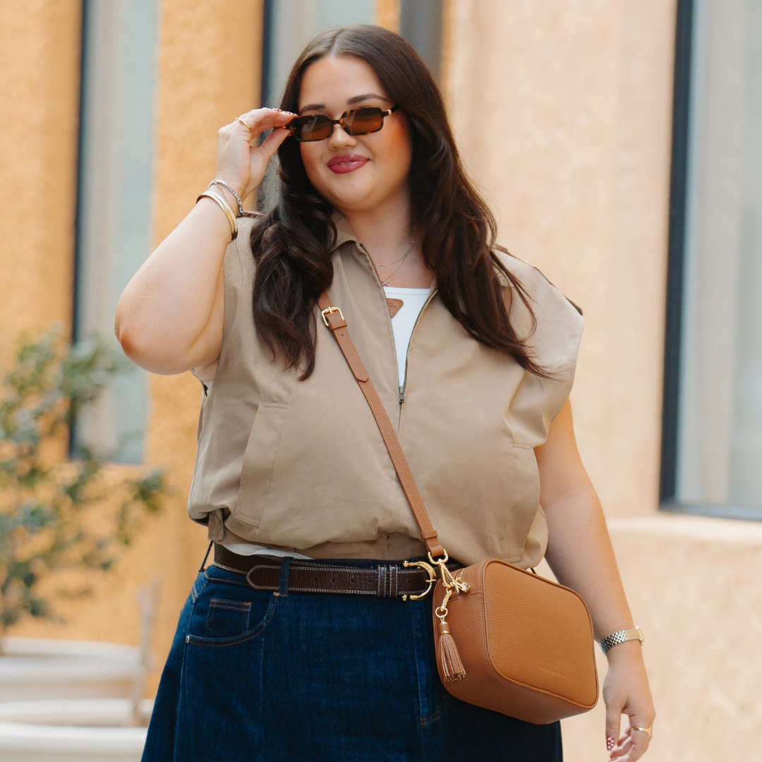 Woman wearing a beige top and blue jeans, holding a brown crossbody bag outdoors.