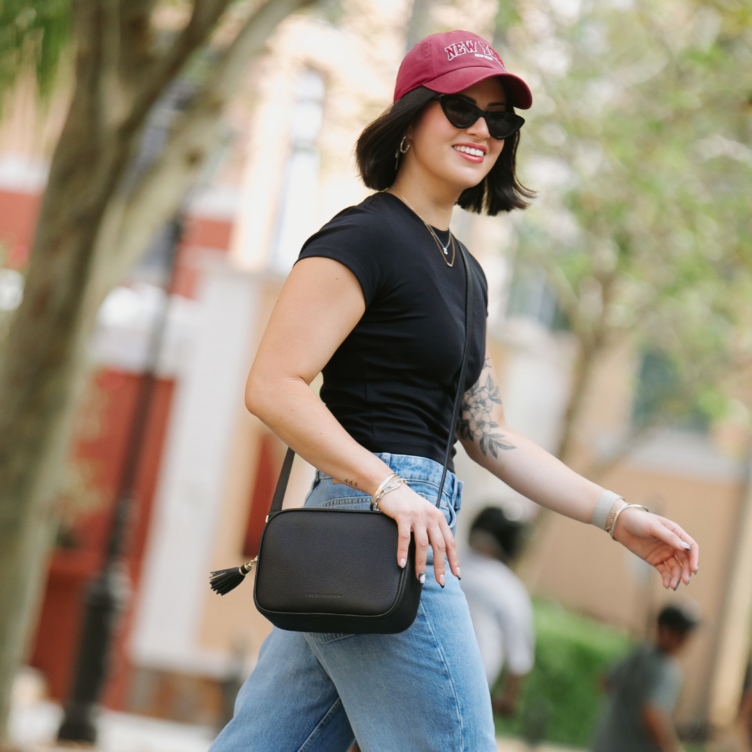 Woman walking outdoors wearing a black top, blue jeans, and a red cap, holding a black leather crossbody handbag.