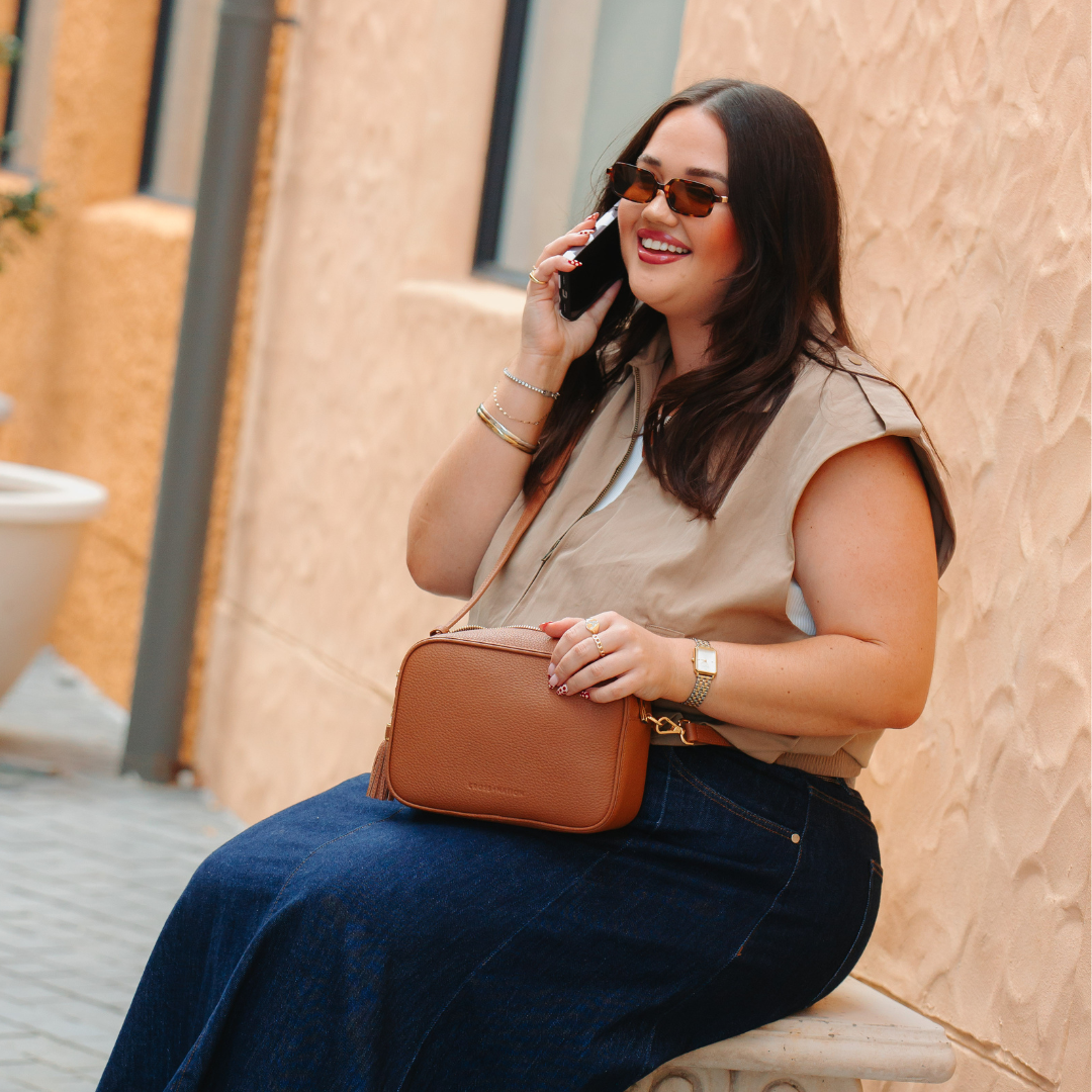 Woman sitting outdoors, talking on a phone with a brown leather crossbody bag, wearing sunglasses and a beige sleeveless top.
